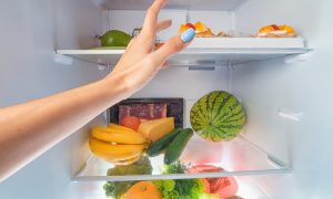 A woman’s hand reaches into an open fridge full of fresh produce such as fruits and other items, some of which may not be part of an elimination diet.